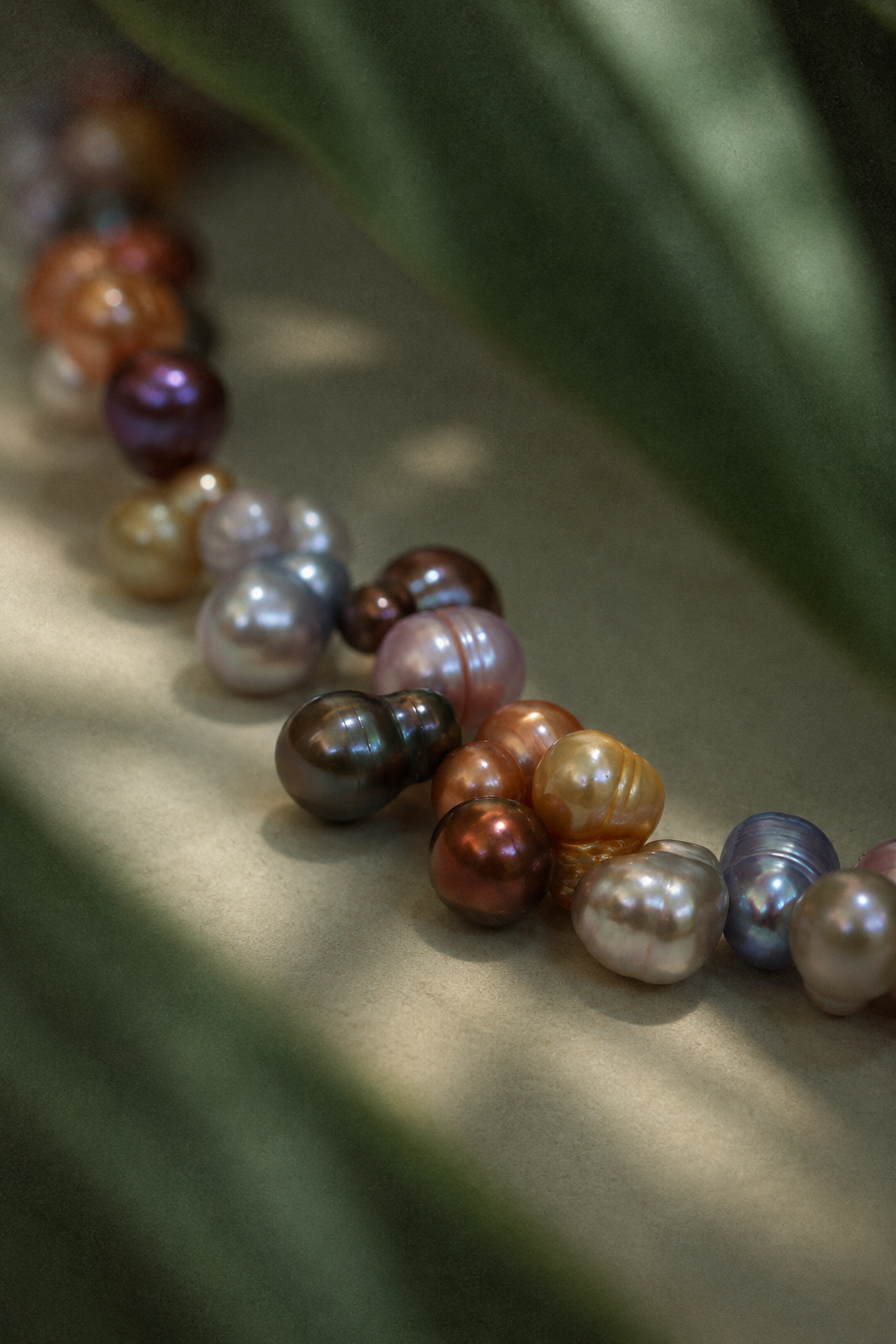 A close-up of a strand of baroque freshwater pearls in various colors, including brown, purple, pink, orange, and white, arranged diagonally on a neutral surface with soft-focus green leaves in the background.